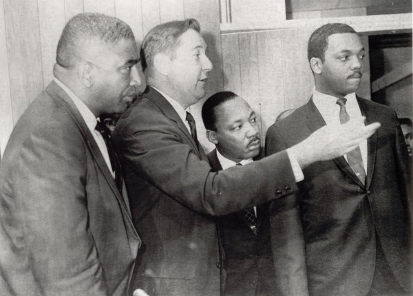 Photographic print of four men in suits standing together indoors, one gesturing with an outstretched arm toward another in the group