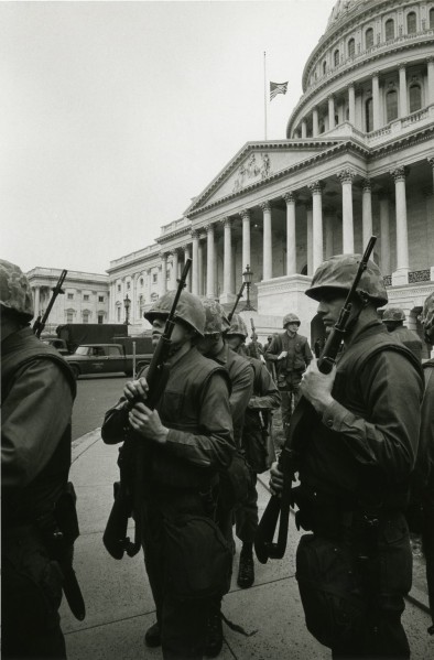 Photographic print of armed, helmeted troops marching in formation in front of the U.S. Capitol building, with a flag flying at half-staff