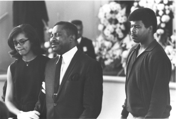 Photographic print of a man in a suit weeping among mourners at a funeral, with large floral arrangements visible in the background