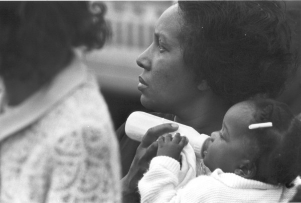 Photographic print of a woman holding a baby with a bottle, gazing solemnly into the distance. Another figure in a patterned coat is partially visible in the foreground