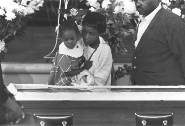 Photographic print of a woman holding a young child as they look into an open casket, surrounded by floral arrangements in a church setting