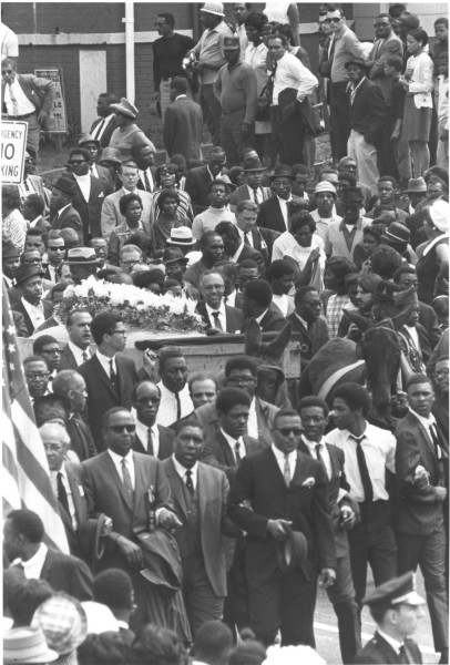 Photographic print of a dense funeral procession moving through city streets, with a flower-draped coffin on a wooden carriage surrounded by thousands of mourners