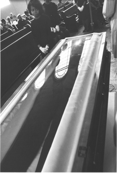 Photographic print of a woman in black standing beside a glass-enclosed coffin in a church, with pews of mourners visible behind her