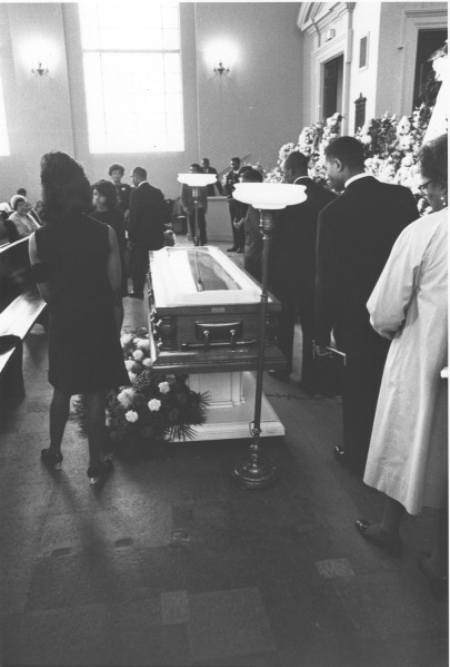 Photographic print of mourners in formal attire walking past an open coffin surrounded by floral arrangements inside a church. Pews, wall sconces, and large windows are visible in the background.