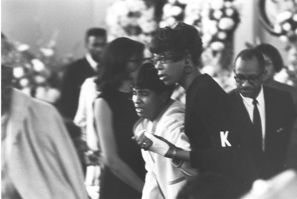 Photographic print showing a distressed woman being supported and guided by another woman indoors, with floral arrangements and a crowd visible in the background