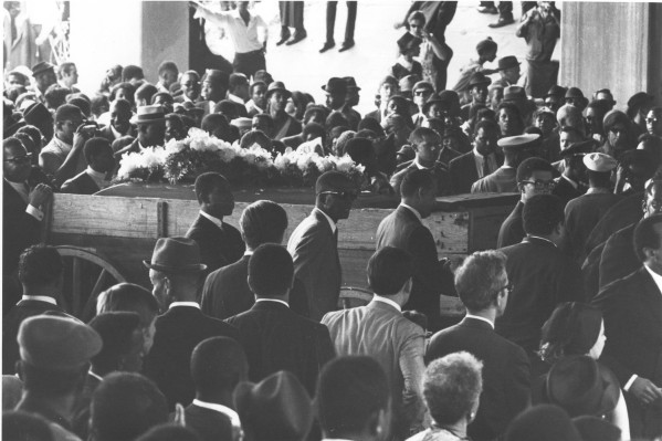 Photographic print of Dr. Martin Luther King, Jr.'s funeral procession. A flower-draped coffin rests on a wooden farm wagon surrounded by a large crowd of mourners.