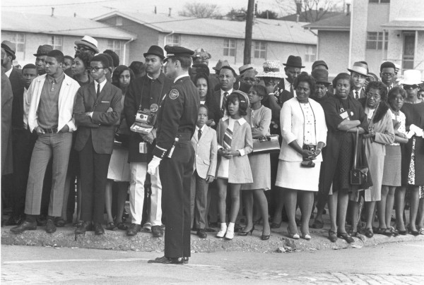 Photographic print of a large crowd of adults and children lining a street, with a uniformed officer standing in the foreground. Residential buildings are visible in the background