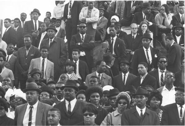 Photographic print of a dense crowd of mourners in formal attire, many wearing suits, ties, and hats, gathered on tiered steps with solemn expressions