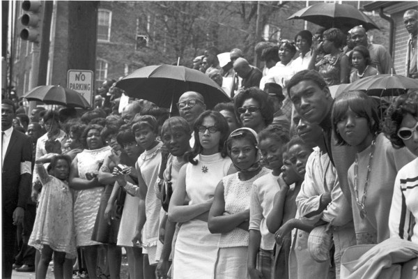 Photographic print of a large crowd of mourners lining an Atlanta street, several holding open umbrellas, with a No Parking sign visible nearby