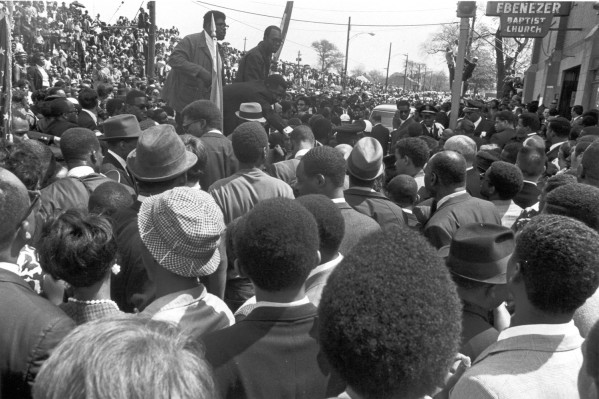 Photographic print of a dense crowd filling the street outside Ebenezer Baptist Church. Two men stand elevated above the crowd, visible against a sign bearing the church's name