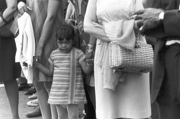 Photographic print of a young girl in a striped dress standing among a crowd of adults on a street, looking directly at the camera