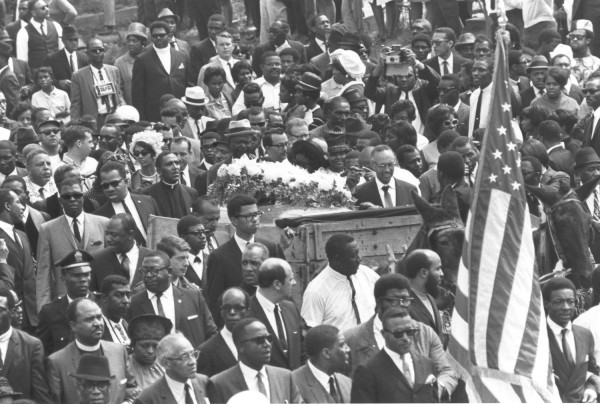Photographic print of Dr. Martin Luther King, Jr.'s funeral procession in Atlanta, with a flower-draped coffin on a wooden carriage surrounded by a dense crowd and an American flag