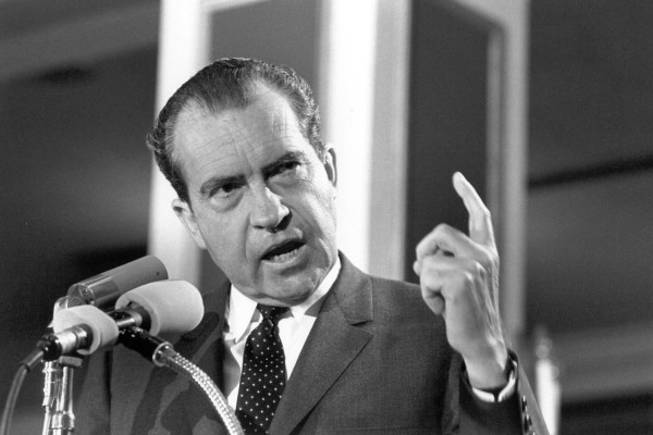 Black-and-white photograph of Richard M. Nixon speaking at a podium with microphones, one finger raised, wearing a dark suit and tie