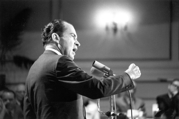 Black-and-white photograph of Richard Nixon speaking at a podium microphone, mouth open, right fist raised. An audience is visible in the background.