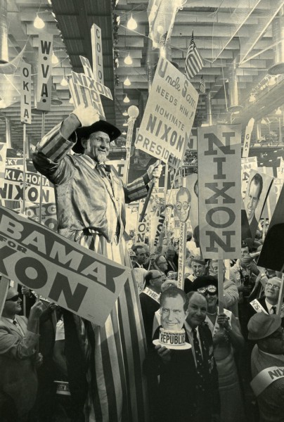 Artifact photograph of a crowded convention floor filled with Nixon campaign signs and state delegation placards, with one attendee dressed in Uncle Sam costume