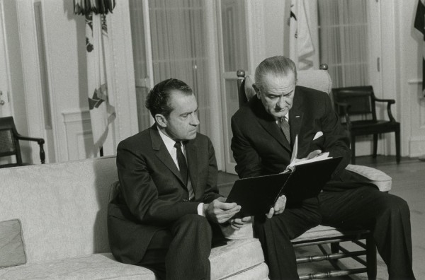 Photographic print of President Johnson and President-elect Nixon seated together in a formal room, reviewing documents held in a dark folder