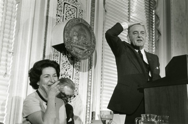 Photographic print of President Johnson gesturing with his hand on his head at a podium, while a smiling woman and another figure sit nearby beneath the National Press Club seal