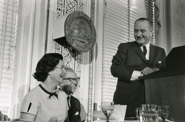 Photographic print of President Johnson laughing at a podium beneath the National Press Club seal, while Lady Bird Johnson and another man seated nearby laugh alongside him
