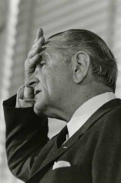 Photographic print of President Johnson in profile, wearing a dark suit, with his right hand raised to his forehead and gazing upward