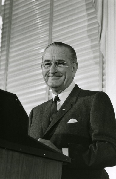 Photographic print of President Lyndon Johnson smiling at a podium, wearing a suit and glasses, with louvered shutters in the background