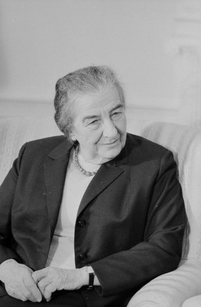 Photographic print portrait of Golda Meir, an older woman seated on a light sofa, wearing a dark blazer and beaded necklace, hands folded