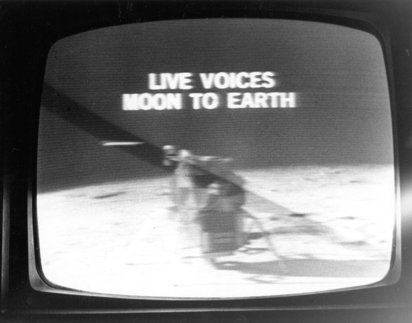 Photographic print of a television screen displaying the text "LIVE VOICES MOON TO EARTH" above a grainy broadcast of a figure on the lunar surface