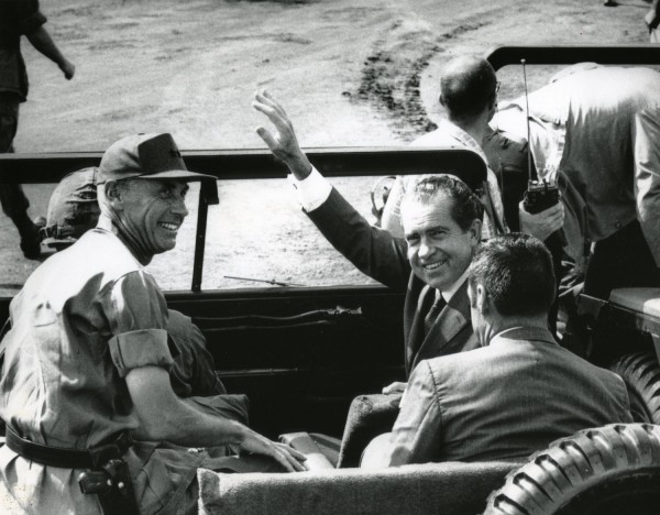 Photographic print of President Richard Nixon waving from an open jeep, seated alongside military officers in uniform