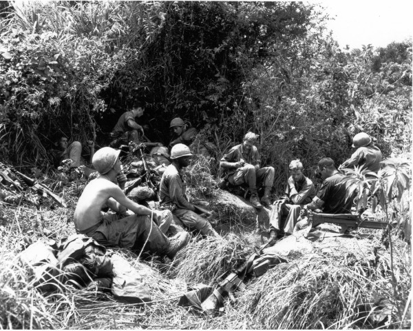 Photographic print of a group of helmeted soldiers resting in tall grass and dense jungle vegetation, with rifles and equipment visible nearby