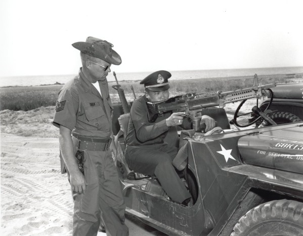 Photographic print of Sergeant Cheek standing beside a military jeep as Captain Tonglee aims an M-60 machine gun mounted on the vehicle, with a coastal landscape in the background