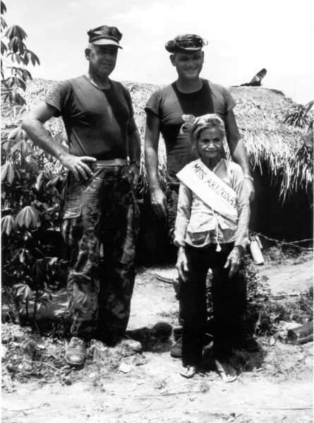 Photographic print of two uniformed Marines standing with an elderly woman wearing a handmade "Miss Arizona" sash, in front of a thatched structure