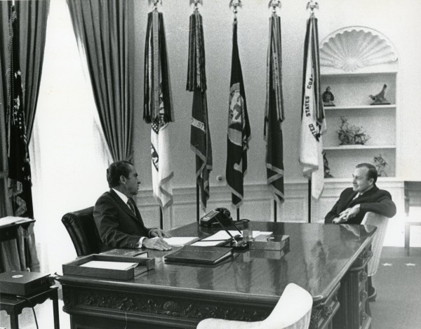 Photographic print of President Nixon seated at the Oval Office desk across from Hugh Sidey, with multiple flags and a bookshelf visible behind them