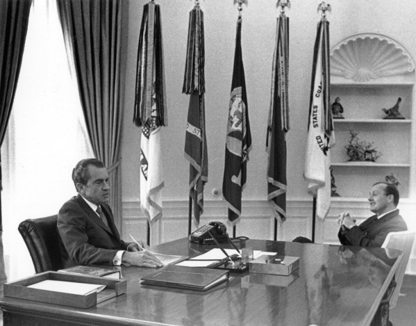 Photographic print of President Nixon and Hugh Sidey seated across a large desk in the Oval Office, with multiple flags displayed behind them