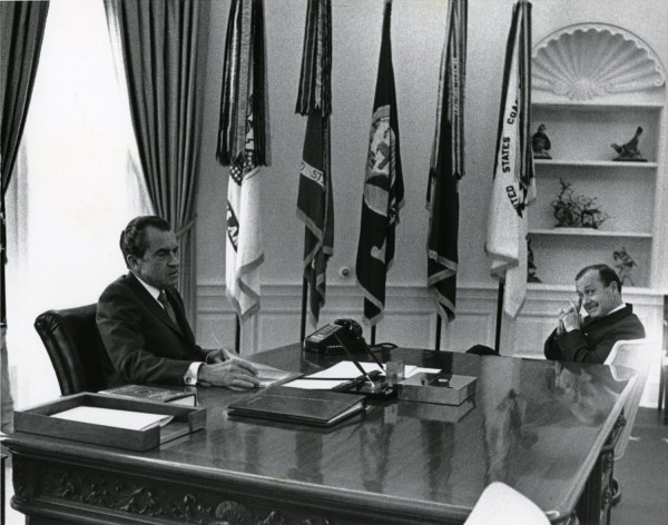 Photographic print of President Richard Nixon seated behind the Oval Office desk across from journalist Hugh Sidey, with several flags and a bookcase visible behind them