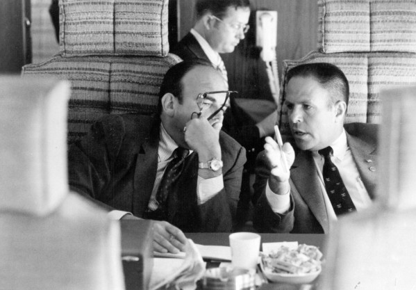 Photographic print of Haldeman and Ehrlichman seated together aboard a plane, conversing at a table with food and drinks nearby
