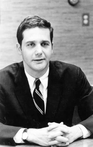 Photographic print of Jeb Magruder seated at a table, hands clasped, wearing a dark suit and striped tie with a watch visible