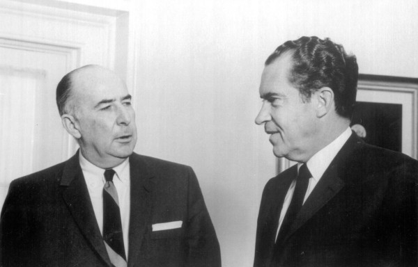 Photographic print of President Nixon and John Mitchell in dark suits and ties, facing each other in conversation against a light wall
