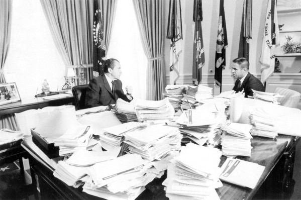 Photographic print of President Richard Nixon and Bob Haldeman seated at a large table covered in tall stacks of papers, with flags and draped windows behind them