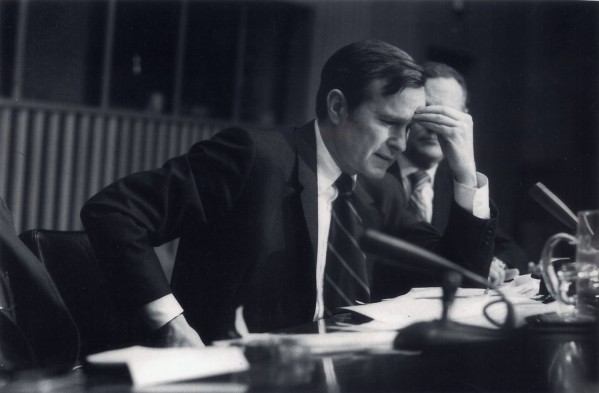 Photographic print of George Bush Sr. seated at a desk with his hand raised to his forehead, papers and a microphone visible in the foreground
