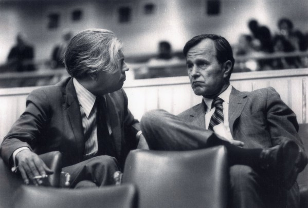 Photographic print of George Bush, with a serious expression, seated in conversation with another suited man in a large assembly hall