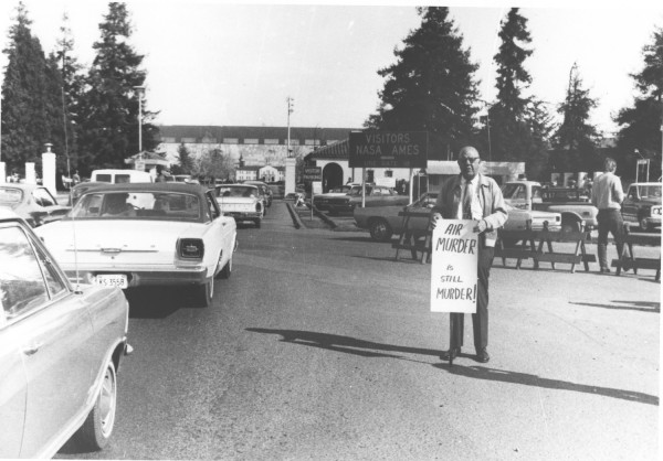 Photographic print of a lone protester standing on a road near the NASA Ames entrance, holding a sign reading "Air Murder is Still Murder!"