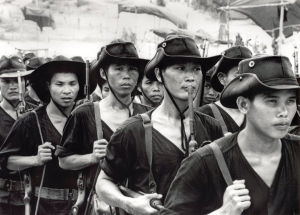 Photographic print of a column of South Vietnamese soldiers in dark uniforms and wide-brimmed hats, carrying rifles with slings over their shoulders
