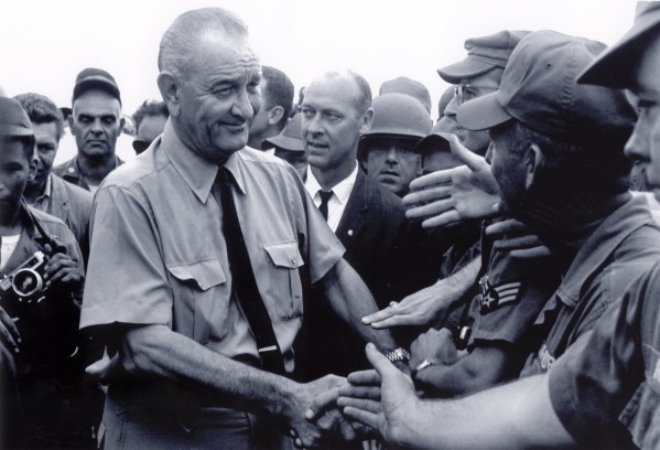 Photographic print of President Lyndon B. Johnson in a short-sleeve shirt and tie shaking hands with uniformed military personnel in caps and helmets, surrounded by a crowd