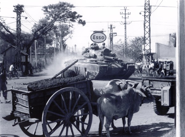 Photographic print of a U.S. Army tank moving down a dusty street past an Esso sign, with an ox-drawn cart in the foreground and bystanders on bicycles nearby