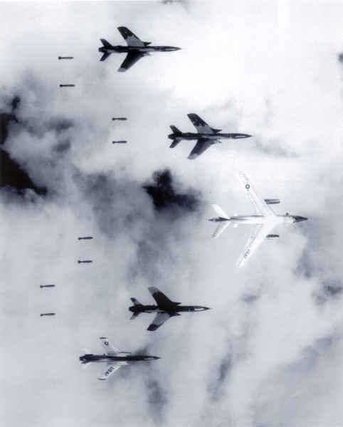 Photographic print of a B-66 Destroyer and four F-105 Thunderchief aircraft in formation, releasing cascading bombs through patchy cloud cover