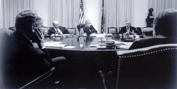 Photographic print of several men in dark suits seated around a large conference table with documents and glasses, an American flag visible in the background