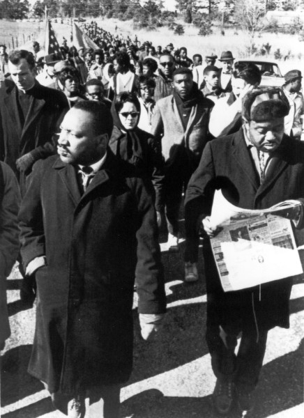 Photographic print showing Dr. Martin Luther King, Jr. and Rev. Ralph Abernathy at the front of a large column of marchers on a rural road, one man carrying a newspaper