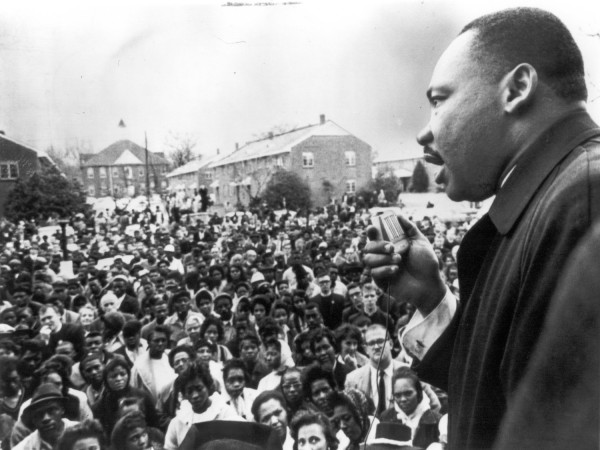 Photographic print of Dr. Martin Luther King, Jr. speaking into a microphone before a large outdoor crowd, with brick buildings visible in the background