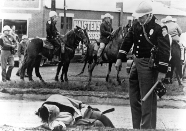 Photographic print of a uniformed officer standing over a woman lying on a street, with mounted police on horseback visible in the background near a brick storefront