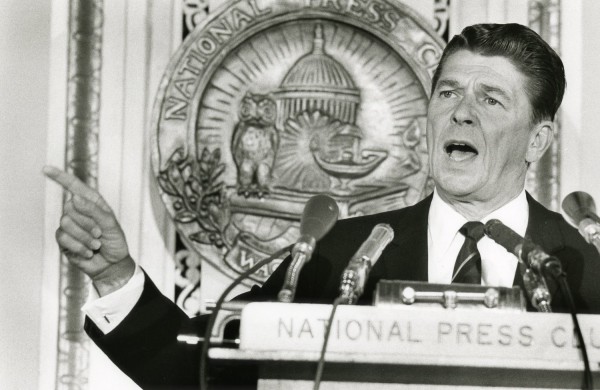 Photographic print of Ronald Reagan speaking at a National Press Club podium with multiple microphones, gesturing with one hand raised