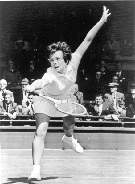 Photographic print of Billie Jean King mid-swing on a tennis court, one arm raised, racket in hand, spectators seated in the background